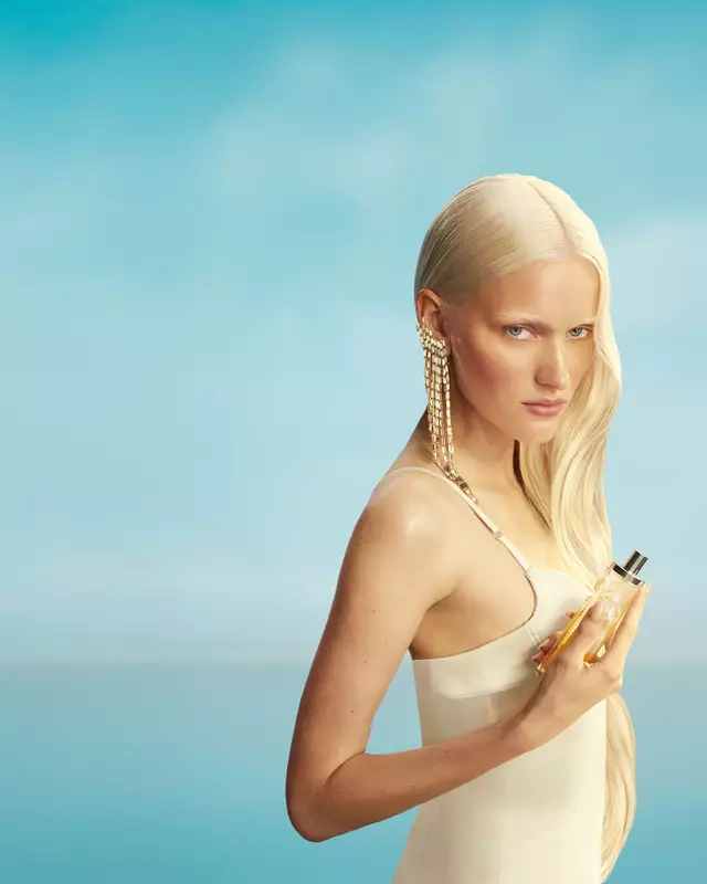 Nouvelle bannière de campagne Mugler Starlicious les eaux gourmandes représentant un mannequin aux longs cheveux blonds et aux yeux bleus, portant une robe jaune pâle et tenant le flacon de parfum Pineapple Musk sur un fond de ciel bleu tendre. Les parfums sont délicieux, à collectionner et à superposer.